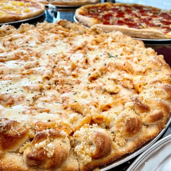 Large pizza with penne pasta, melted cheese, and a garlic knot crust, with other pizzas in background.