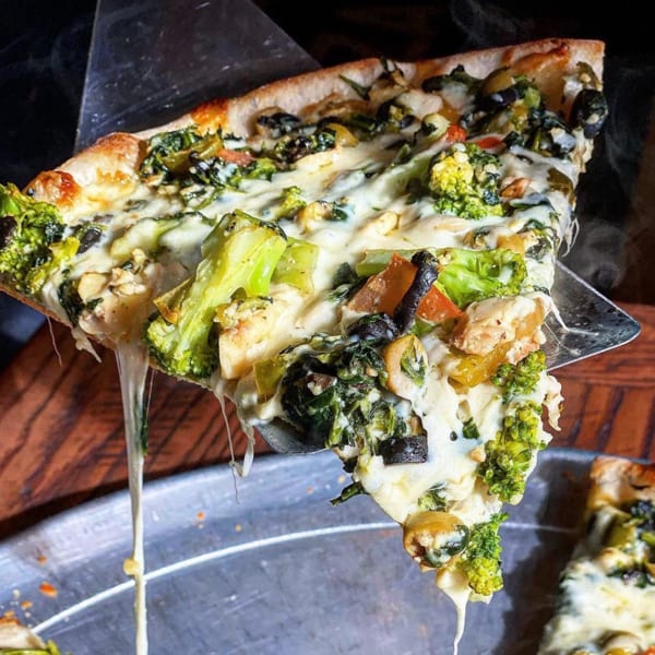 A pizza slice with broccoli, olives, and melted cheese stretching from a spatula.