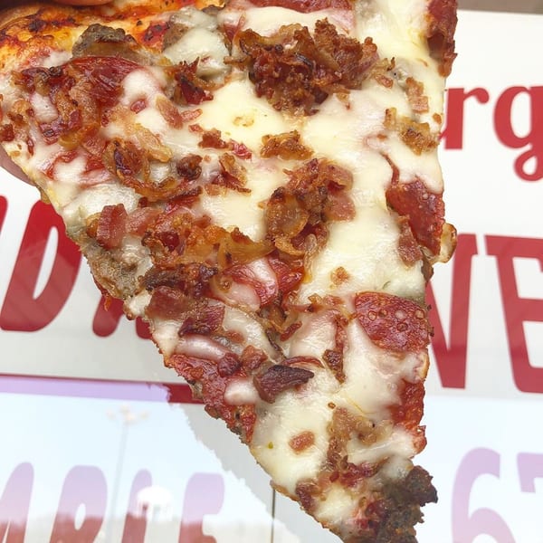 Hand holding a pizza slice loaded with pepperoni, bacon, and melted cheese.