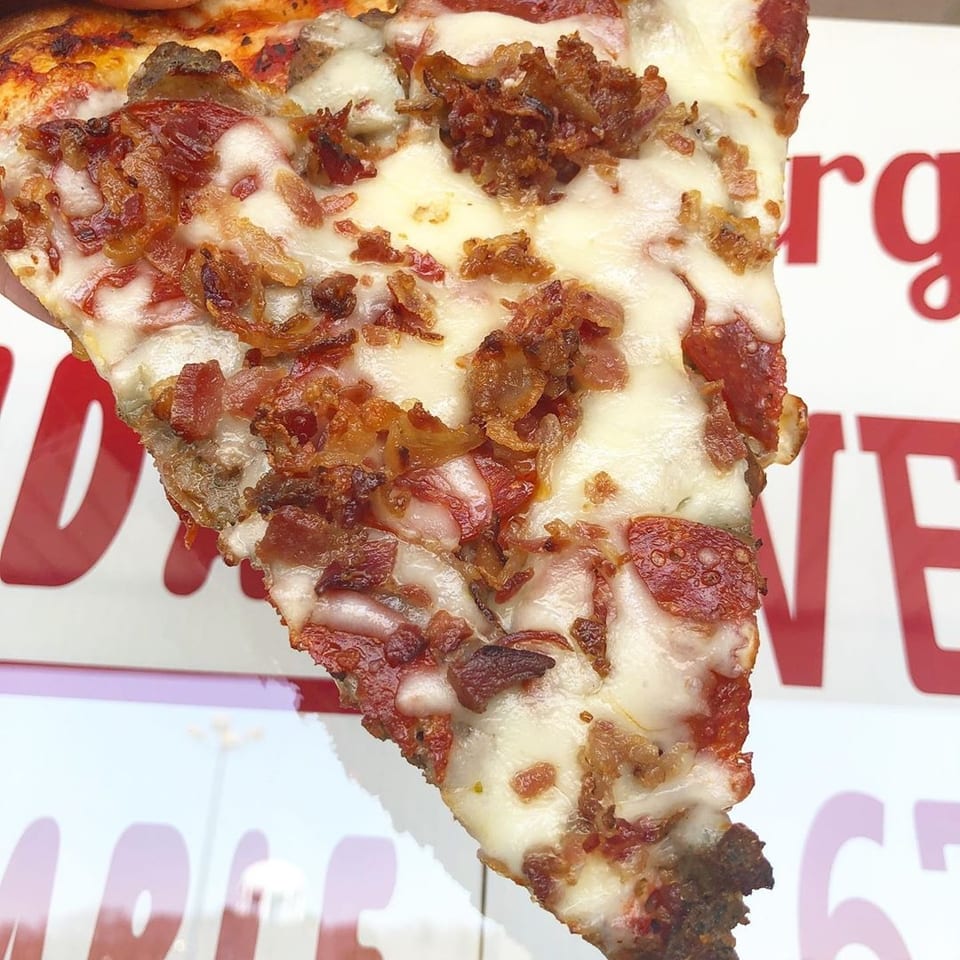 Hand holding a pizza slice loaded with pepperoni, bacon, and melted cheese.